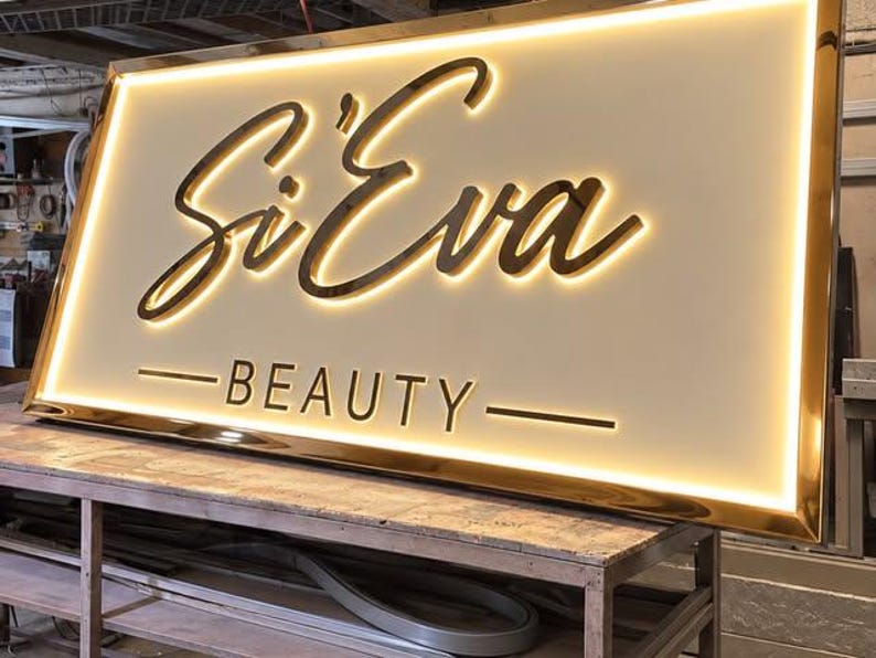 May include: A rectangular, wall-mounted sign with a gold-toned metallic frame and a white face. The sign features the text Si'Eva in a large, dark, cursive script and the word BEAUTY in a smaller, sans-serif font, both illuminated by backlighting.