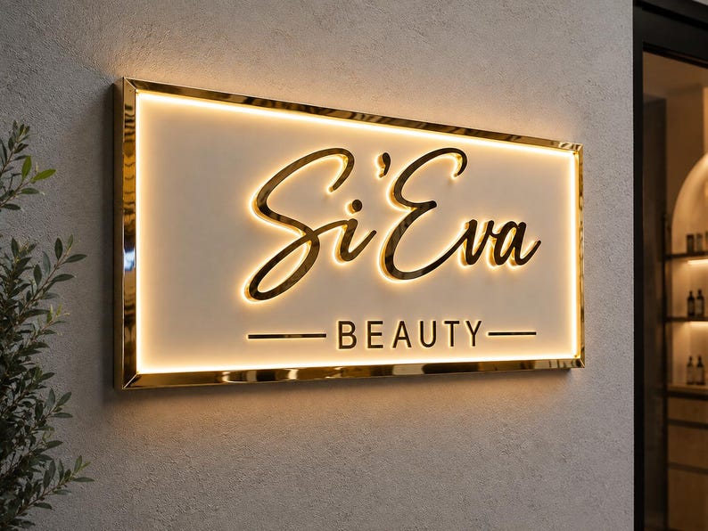 May include: A rectangular wall-mounted sign with a gold metallic frame and a white face. The sign features the text Si'Eva in a gold cursive script and the word BEAUTY in gold block letters, both illuminated by a warm internal LED glow.