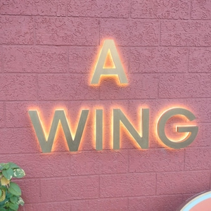 May include: A wall-mounted sign featuring the text A WING in large, metallic gold-toned letters. Each letter is backlit with a warm, glowing orange light, creating a halo effect against the textured, reddish-brown brick surface of the wall.