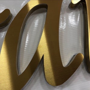 May include: A three-dimensional cursive letter sign featuring a brushed gold metallic finish on the front surface. The letter has a thick profile with a clear acrylic layer visible along the edges, creating a layered effect with depth and dimension.