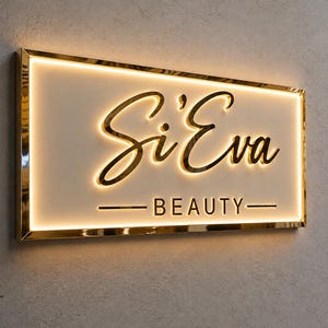 May include: A rectangular wall-mounted sign with a gold metallic frame and a white face. The sign features the text Si'Eva in a gold cursive script and the word BEAUTY in gold block letters, both illuminated by a warm internal LED glow.