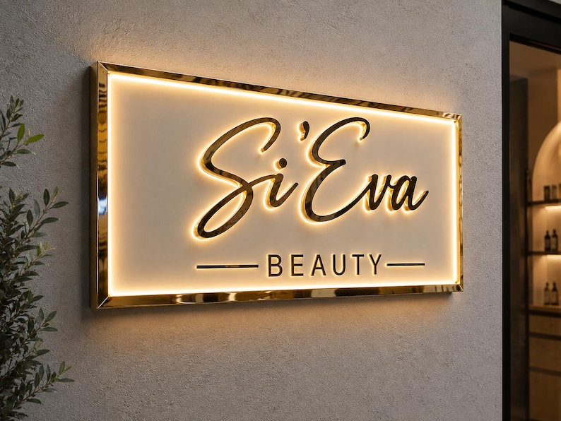 May include: A rectangular wall-mounted sign with a gold metallic frame and a white face. The sign features the text Si'Eva in a gold cursive script and the word BEAUTY in gold block letters, both illuminated by a warm internal LED glow.