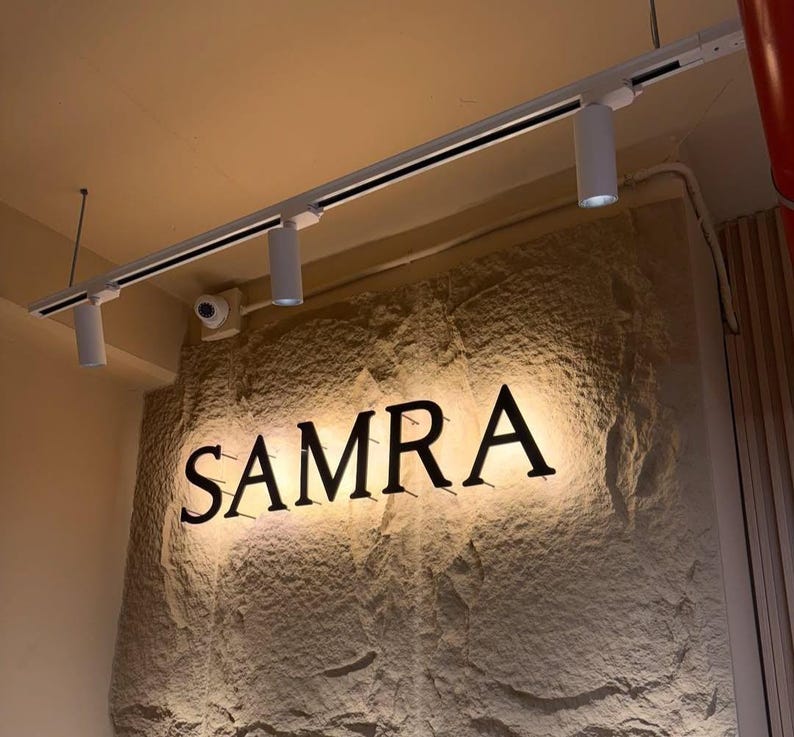 May include: A wall-mounted sign featuring the black capital letters SAMRA mounted on a textured, light-colored stone surface. The sign is illuminated by a white track lighting system with three cylindrical spotlights attached to the ceiling above.
