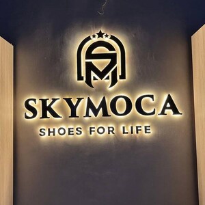 May include: A wall-mounted sign featuring the black text SKYMOCA and the slogan SHOES FOR LIFE, positioned below a stylized logo with three stars. The letters and logo are backlit with a warm glow, creating a halo effect against the dark wall.