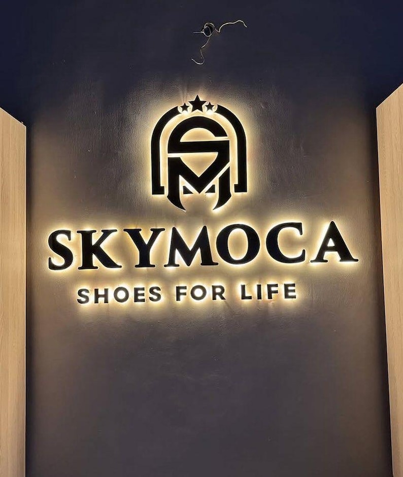 May include: A wall-mounted sign featuring the black text SKYMOCA and the slogan SHOES FOR LIFE, positioned below a stylized logo with three stars. The letters and logo are backlit with a warm glow, creating a halo effect against the dark wall.