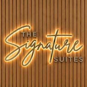 May include: A wall-mounted sign featuring the text THE SIGNATURE SUITES in a combination of block and script lettering. The word Signature is illuminated with a warm golden glow, set against a vertical brown wood slat paneling background.