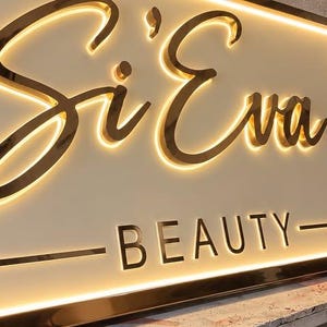 May include: A rectangular wall sign featuring the text Si'Eva in a large, gold-toned cursive script and the word BEAUTY in smaller, gold-toned block letters. The sign has a cream-colored background and is illuminated by a warm, glowing backlight.