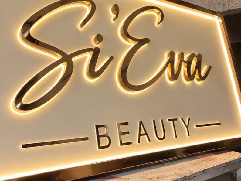 May include: A rectangular wall sign featuring the text Si'Eva in a large, gold-toned cursive script and the word BEAUTY in smaller, gold-toned block letters. The sign has a cream-colored background and is illuminated by a warm, glowing backlight.