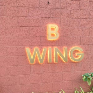 May include: A wall-mounted sign featuring the text B WING in large, gold-colored, three-dimensional letters. The letters are backlit with a warm orange glow, creating a halo effect against the textured, salmon-colored brick wall surface.