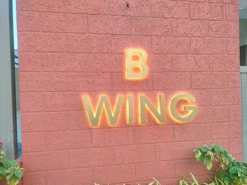 May include: A wall-mounted sign featuring the text B WING in large, gold-colored, three-dimensional letters. The letters are backlit with a warm orange glow, creating a halo effect against the textured, salmon-colored brick wall surface.