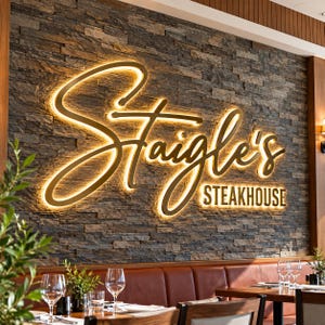 May include: A large, illuminated wall sign featuring the text Staigle's STEAKHOUSE in a gold, cursive script and block lettering. The sign is mounted on a textured, dark gray stone wall, with warm backlighting creating a glowing effect.