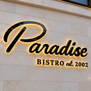 May include: A wall-mounted sign featuring the word Paradise in a large black script font, with the text BISTRO est. 2002 in a smaller black sans-serif font below. Both lines of text are backlit with a warm glow against a light stone wall.