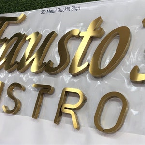 May include: A 3D metal backlit sign featuring the gold-colored text Fausto's BISTRO in a combination of script and block lettering. The letters have a metallic finish and are mounted on a white surface with the text 3D Metal Backlit Sign.