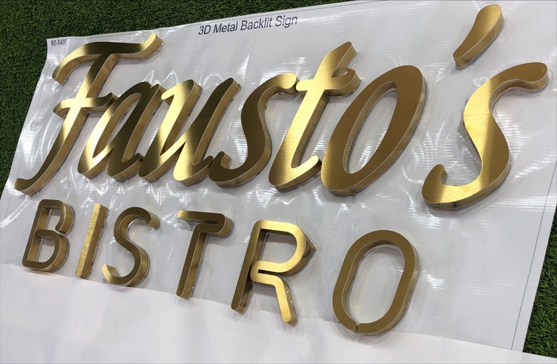 May include: A 3D metal backlit sign featuring the gold-colored text Fausto's BISTRO in a combination of script and block lettering. The letters have a metallic finish and are mounted on a white surface with the text 3D Metal Backlit Sign.