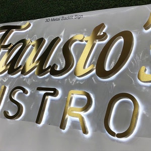 May include: A 3D metal backlit sign featuring the gold-colored text Fausto's BISTRO in a combination of script and block lettering. The letters are mounted on a white surface and emit a bright white glow from behind each individual character.