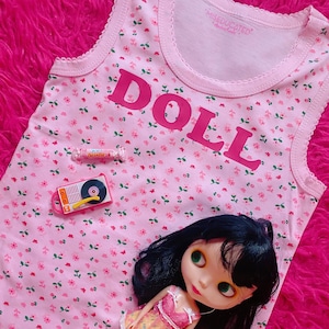 Könnte beinhalten: Ein rosa ärmelloses Tanktop mit Blumenmuster und dem Wort "DOLL" in rosa Buchstaben. Eine kleine Puppe in einem gelben und rosa Kleid liegt auf dem Tanktop.