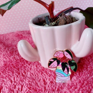 May include: A pink enamel pin featuring a cartoon plant with rainbow stripes and the text "Plant Lover".