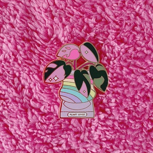 May include: A pink and green enamel pin featuring a plant in a pot with a rainbow design. The pot has the text "PLANT LOVER" on the front.