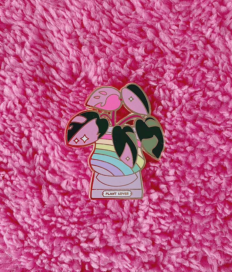 May include: A pink and green enamel pin featuring a plant in a pot with a rainbow design. The pot has the text "PLANT LOVER" on the front.