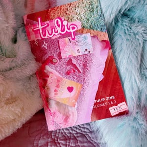 Könnte beinhalten: Ein Exemplar von "Tulip Zine Volumes 1 & 2" mit einem rosa-weißen Cover, auf dem das Wort "tulip" in Schreibschrift steht. Das Zine wird von zwei kleinen Karten begleitet und liegt auf einer rosa gesteppten Oberfläche, umgeben von flauschigen Textilien.