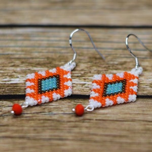 Puede incluir: Un par de pendientes de cuentas naranja, blanco y azul con ganchos de plata. Los pendientes tienen forma de diamante y tienen una pequeña cuenta roja en la parte inferior.