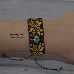 May include: A beaded bracelet with a black background and yellow, red, blue, and white geometric patterns. The bracelet has a black cord with yellow and blue beads at the ends.