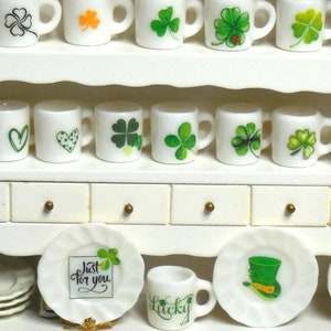 Puede incluir: Tazas de cerámica blanca con diseños de trébol y corazón en un aparador blanco con tres cajones. Las tazas están dispuestas en filas en los estantes. También hay platos de cerámica blanca con diseños de trébol y duendes en el estante inferior.