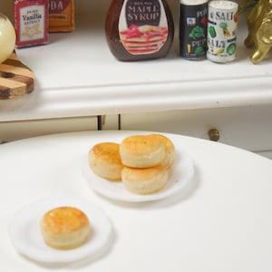 May include: Miniature breakfast scene featuring a white table with two small plates of scones. A small pitcher of liquid, maple syrup, and spices are on a white shelf in the background. A gold clock and red roses are also visible.