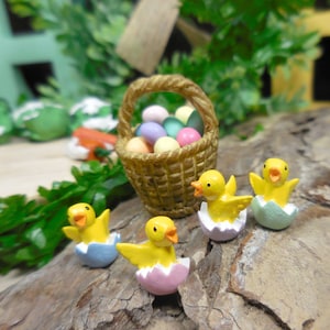 May include: A miniature Easter scene featuring a woven basket filled with pastel-colored eggs. Several yellow chicks are emerging from pastel-colored eggshells. The scene is set on a piece of wood, with greenery in the background.