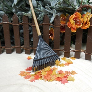 Puede incluir: Un rastrillo de metal negro con mango de madera se apoya en una valla de madera marrón.  Hojas de otoño de color naranja y amarillo están esparcidas en el suelo frente al rastrillo.