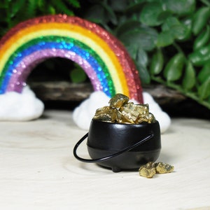 May include: A black pot filled with gold nuggets sits on a wooden surface, with a rainbow and clouds in the background. The pot has a handle and is surrounded by green foliage, evoking a St. Patrick's Day theme.