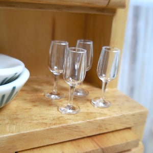 Puede incluir: Cuatro copas de champán de plástico transparente en miniatura sobre una superficie de madera. Las copas son delgadas con un diseño clásico, perfectas para una casa de muñecas. Un cuenco blanco y verde es visible.