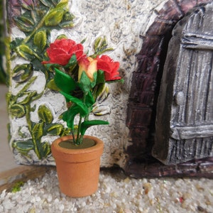 May include: A miniature terracotta pot with a small rose plant. The plant features red and yellow roses with green leaves. The pot is set against a textured, white and green background, near a miniature door.