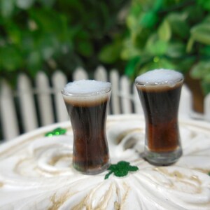 May include: Two miniature glasses filled with dark brown liquid topped with white foam. The glasses are on a white surface with green shamrocks.