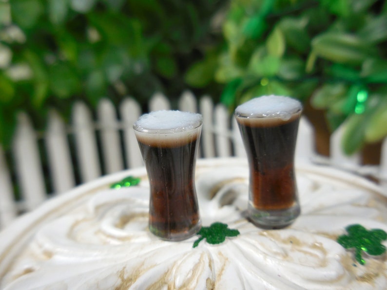 May include: Two miniature glasses filled with dark brown liquid topped with white foam. The glasses are on a white surface with green shamrocks.