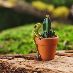 Pode incluir: Uma figurinha de sapo verde empoleirada na borda de um vaso de terracota, que contém uma pequena planta de cacto com uma única flor rosa.