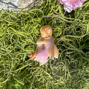 Può includere: Una piccola statuina di fata in porcellana con ali rosa e un vestito rosa. La fata è seduta su un letto di muschio verde.