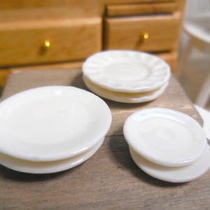 Puede incluir: Tres juegos de platos de cerámica blanca, apilados en diferentes tamaños, sobre una superficie de madera. Los platos son todos redondos y tienen un acabado liso y brillante.