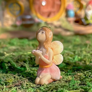 May include: A small, pink fairy figurine with a flower crown and wings. The fairy is holding a small flower in her hands and is kneeling on a bed of green moss.