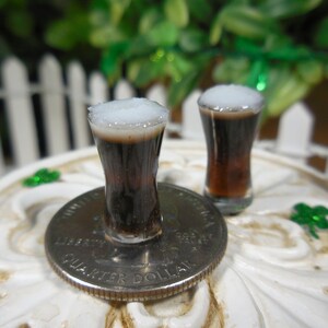 May include: Two miniature glasses of dark beer with white foam on top, sitting on a white surface with a silver US quarter dollar coin.
