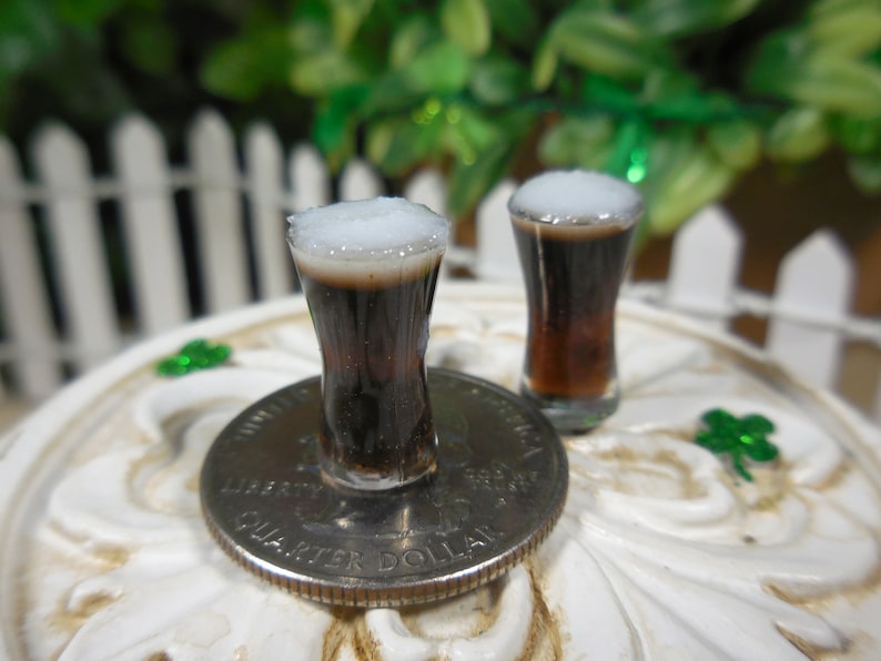 May include: Two miniature glasses of dark beer with white foam on top, sitting on a white surface with a silver US quarter dollar coin.