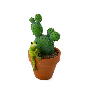 Puede incluir: Una figurita de rana verde posada en el borde de una maceta de terracota, con una planta de cactus verde en su interior. El cactus tiene una forma redonda y espinosa y está plantado en una maceta marrón.