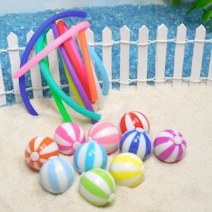 Puede incluir: Un conjunto de nueve pelotas de playa de colores con rayas blancas, dispuestas sobre una superficie de arena blanca frente a una valla blanca de madera. Las pelotas de playa son de plástico y tienen un acabado brillante.