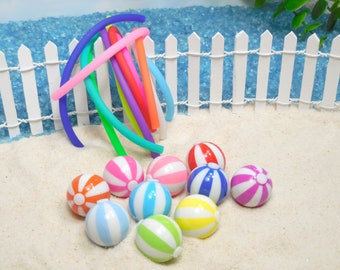 May include: A set of nine colorful beach balls with white stripes, arranged on a white sandy surface in front of a white picket fence. The beach balls are made of plastic and have a glossy finish.
