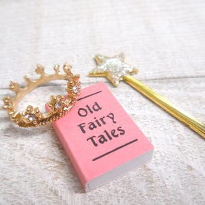 Może przedstawiać: Złoty pierścionek w kształcie korony z przezroczystymi kamieniami, różowa książka z napisem "Old Fairy Tales" i złota różdżka z gwiazdą na końcu. Przedmioty ułożone są na białej, teksturowanej powierzchni, nawiązując do tematu bajki.