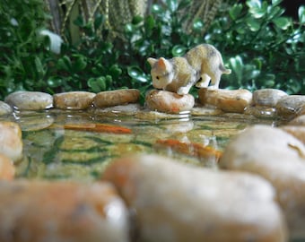 May include: A miniature white cat figurine with brown markings is perched on rocks surrounding a small pond with clear water and two orange goldfish. The pond is surrounded by green foliage.
