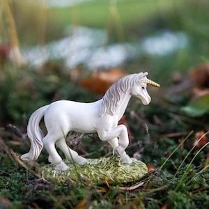 Puede incluir: Una figura de unicornio blanco con un cuerno dorado y una melena fluida se alza sobre un trozo de hierba verde. La criatura mítica está posada en una postura dinámica, sobre un fondo borroso de vegetación.