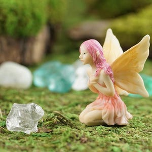 May include: A small, pink and yellow fairy figurine with wings, kneeling on a bed of green moss. The fairy has long pink hair and is wearing a pink dress.