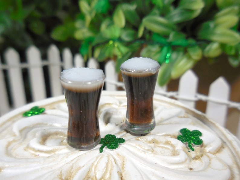 May include: Two miniature glasses of dark brown liquid with white foam on top. The glasses are on a white surface with green shamrock decorations.