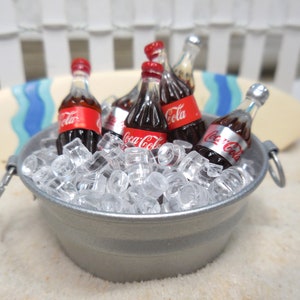 May include: A silver metal bucket filled with ice and miniature Coca-Cola bottles. The bucket is sitting on a white sandy surface.